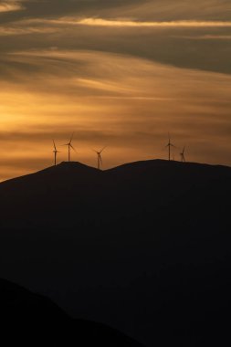 Renkli gökyüzünde gün doğumu, günbatımı. Altın güneş ışınları ufku boyuyor, tepelerdeki rüzgar jeneratörleri enerji veriyor. Bulutların arasından geçen güneş, dağların üzerinden geçen siluet, arka plan bulutları. Dikey fotoğraf.