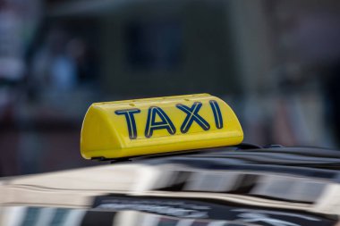 Arabanın tavanında taksi tabelası, sarı ve siyah renkli metin, yakın çekim görüntüsü, gündüz vakti şehirde taksi, yolcu bekliyor, şehir manzarası bulanık.