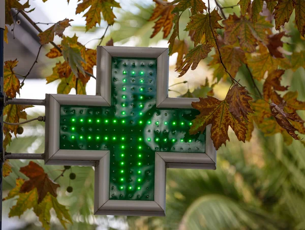 Green cross pharmacy illuminated sign hanging outside drugstore, tree foliage background