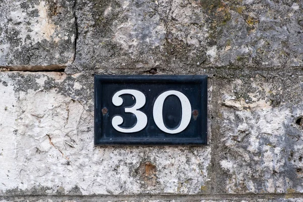 Street number thirty. Digit 30 white color on metal vintage plate mounted on stone wall. Traditional house address