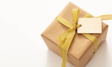 Beyaz arka planda izole edilmiş bir hediye kutusunun üzerindeki boş etiket, yakın plan görüntüsü. Kraft geri dönüşümlü kahverengi kağıtlı, altın sarısı kurdeleli, Noel hediyesi şablonu, kopyalama alanı