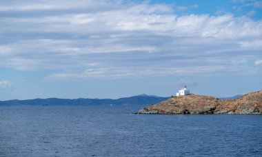 Yunanistan, Kea Tzia adası. Kayalık bir pelerin üzerindeki beyaz deniz feneri, bulutlu mavi gökyüzü ve dalgalı koyu renkli deniz suyu arka planı. Yaz, turist beldesi. Hava aracı görünümü