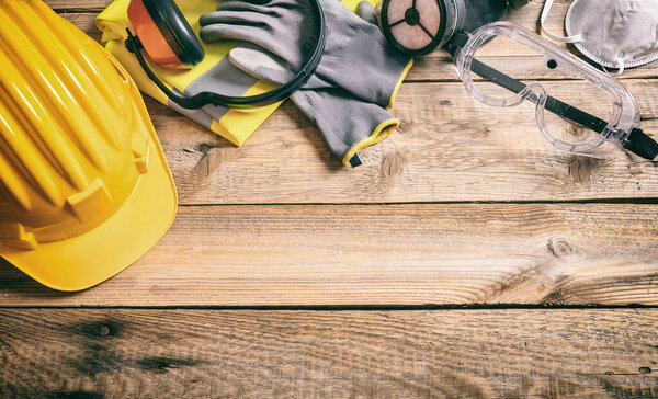 Construction work safety concept. Protective yellow hard helmet, headphones, gloves, work vest, mask and goggles on wooden background. Protection and prevention equipment for workers. Banner, space.