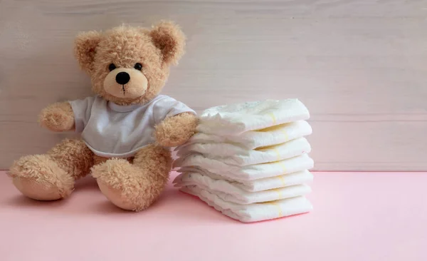 Baby diapers stack and cute teddy bear sitting on pink color floor, wooden wall background. Newborn girl nappies concept