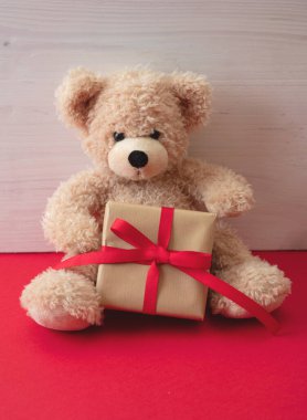 Sevgililer günü, Noel hediyesi. Kırmızı zeminde duran hediye kutusunu tutan oyuncak ayı, tahta duvar arkaplanı, fotokopi alanı, doğum günü tebrik kartı