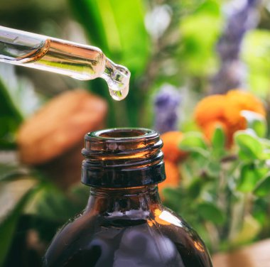 Alternatif tıbbi, homeopati, aromaterapi, pipetten şişeye gerekli yağ damlası. Düşürücü arka planda bulanık bitki çeşitliliğine yakın plan.