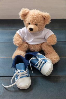 Küçük beden ayakkabılar, bez ayakkabılar bağcıksız ayakkabı bağcığı ile mavi-beyaz paçalar. Bebek spor ayakkabıları ve mavi ahşap zeminde sevimli bir oyuncak ayı. Çocuk odasının içi