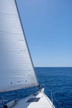 White sloop rigged yacht sailing in open calm ocean, blue sky background, sunny day. Sailboat deck, bow, mast and sails closeup view. Leisure activity, travel and transportation concept.