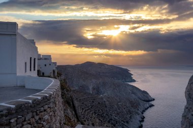 Folegandros island at sunset. Cyclades, Greece. Breathtaking view over the Aegean sea from high cliffs at chora village, Sun shine on orange color cloudy sky, calm rippled sea water