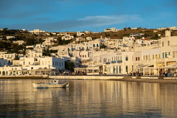 Mykonos island, Cyclades, Greece. May 20, 2021. Wooden fishing boat moored, old harbor cityscape at sunset light, houses shops, reflections on calm Aegean sea. Greek famous destination luxury resort.