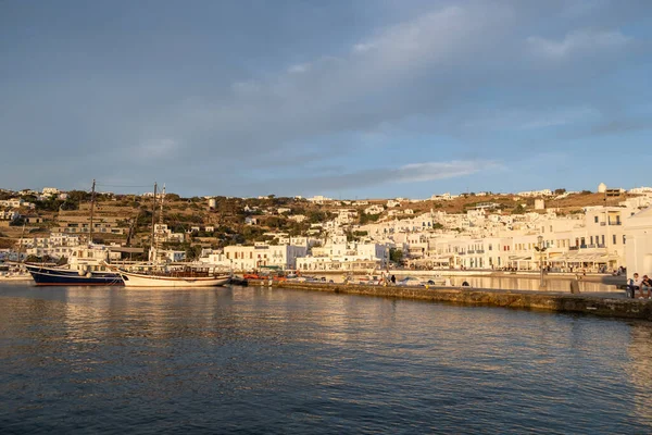 Mykonos island, Cyclades, Greece. May 20, 2021. Old port cityscape at sunset light, houses shops and boats, calm Aegean sea, cloudy blue sky background. Greek famous destination luxury resort.