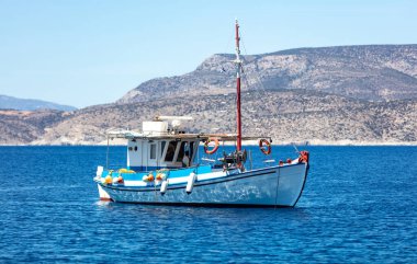 Açık denize demirlemiş renkli bir balıkçı teknesi, lo adasının arka planında. Cyclades, Yunanistan. Geleneksel eski ahşap gemi durgun sulara demirlemiş. Güneşli gün, açık mavi gökyüzü