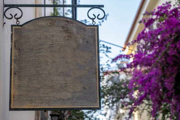 Blank empty wood sign hanging of a metal structure, blue sky and blooming bougainvillea, Nafplio old town narrow alley, Greece. Copy space, advertise template