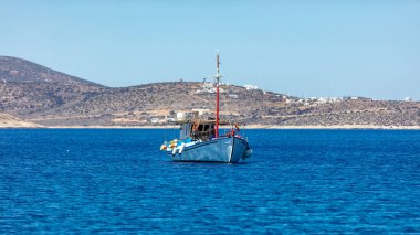 Açık denize demirlemiş renkli bir balıkçı teknesi, lo adasının arka planında. Cyclades, Yunanistan. Geleneksel eski ahşap gemi durgun sulara demirlemiş. Güneşli gün, açık mavi gökyüzü