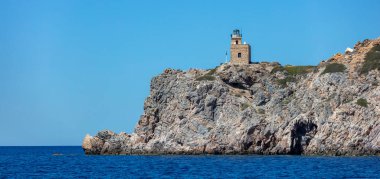 Kayalık bir uçurumda deniz feneri binası. Yunanistan adası. Limanın girişinde bir pelerin üzerinde taş fener, açık mavi gökyüzü arka planı, sakin deniz.