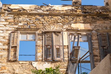 Hasarlı taş duvar binası bozulmuş eski ahşap panjurlar ve mavi gökyüzü manzaralı pencereler. Terk edilmiş eski çürümüş ev, Syros Adası Cyclades Yunanistan 'daki terk edilmiş yapıyı mahvetti. Güneşli yaz günü.