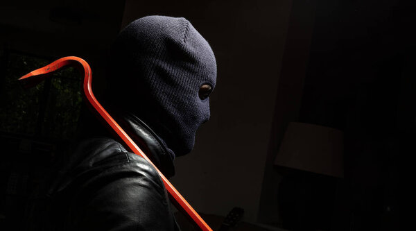 Hooligan in balaclava with red crowbar, in dark night background. Masked burglar holding a lever, weapon and tool for vandalism and violence.