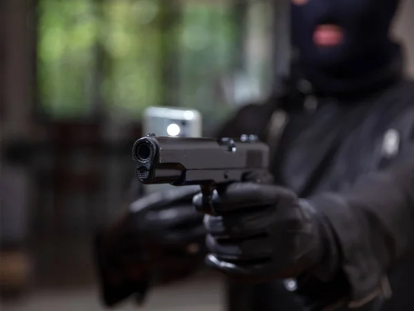 Videotaping your death concept. Blur terrorist in black balaclava holding pistol in leather gloved one hand and mobile in the other. Armed man killer robber murderer aims with pistol before shooting.