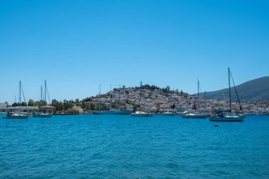 Poros Adası Yunanistan Limanı. Sakin mavi sularda yüzen tekneler, geleneksel mimari tepe evleri, ahşap iskele ve yaz gökyüzü.