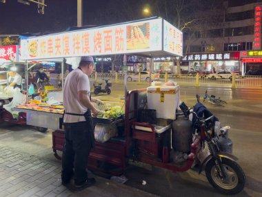 Xian, Çin, 26 Mart 2025. Bir gece yemeği satıcısı yol kenarındaki tezgahta şiş hazırlıyor, renkli tabelalar ve geçen trafik işaretleri.