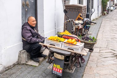 Suzhou, Çin, 30 Mart 2025. Sokak satıcısı eski dar bir kasaba sokağında portakal, biber ve üzümlerle dolu bir meyve arabasının yanında oturur.
