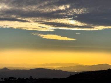 Atina, Yunanistan 'ı çevreleyen tepelerin üzerinde ılık altın gün batımı, yumuşak akşam ışığında parlayan katmanlı bulutlar ve sakin bir gökyüzü altında sisli ufukta kaybolan uzak dağlar..