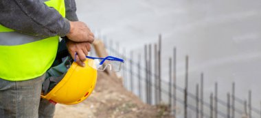 İnşaat alanında çelik yelek ve gözlük tutan işçiler beton ve demir parmaklıkların yanında..