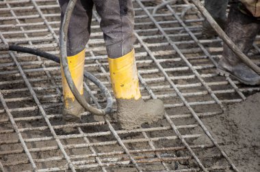 Sarı çizmeli işçiler inşaat alanındaki çelik takviye şebekesinin üzerine ıslak beton döküyorlar. Gün ışığında yakın çekim görülüyorlar..