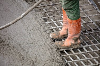 İnşaat işçisi çelik parmaklıkların üzerine ıslak beton döküyor, turuncu çizmeler ve yeşil pantolon giyiyor, bulutlu bir günde yukarıdan görünüyor..