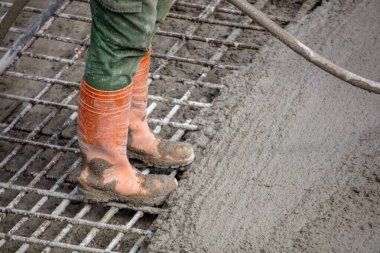 Turuncu çizmeli işçi inşaat alanındaki çelik inşaat ızgarasının üzerine ıslak beton döküyor..