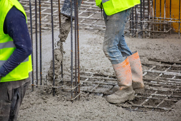 Работники в защитных жилетах и сапогах заливают мокрым бетоном укрепленный фундамент на строительной площадке, захваченный при дневном свете с видимыми стальными стержнями и мутной текстурой.