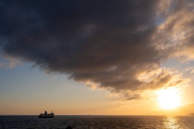 Feribot sakin denizi aşar dramatik günbatımı gökyüzünün altında, kara bulutlar suyun üzerinde yavaşça parlar