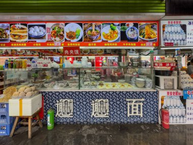 Xian, Çin, 27 Mart 2025. Gıda pazarındaki geleneksel bir erişte tezgahında tabaklar, tabaklar ve içecekler parlak tabelalar altında sergileniyor.