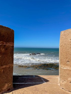 Eski Essaouira surları arasında çerçevelenmiş Atlantik dalgaları, taş tahkimatı, canlı mavi gökyüzü, güneşli gün ve sakin okyanus manzarası