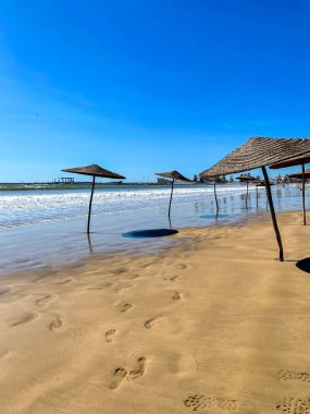 Fas 'ın güneşli Essaouira kıyı şeridi boyunca ıslak kumsalda boş plaj şemsiyeleri, mavi gökyüzünün altında süzülen hafif dalgalar.