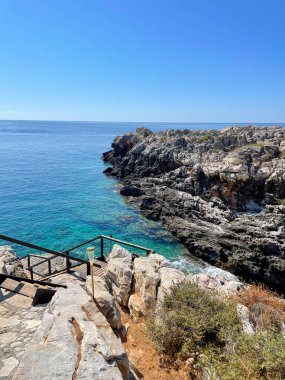 Gün ortası güneşi altında turkuaz suların üzerindeki kayalık kıyı terası, sakin Ege Denizi, Girit Adası, Yunanistan 'a inen ahşap basamaklar