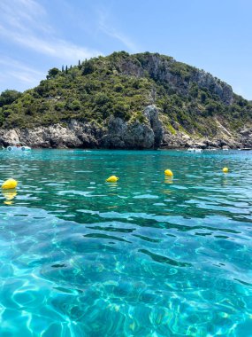 Korfu, Yunanistan 'da turkuaz deniz suyuyla kayalık kıyı şeridi. Güneşli bir yaz günü, berrak gökyüzü, yemyeşil ve sudaki sakin yansımalar