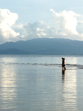 Koh Samui, Tayland, 6 Kasım 2025. İki insan kıyı kenarında sığ deniz suyunda yürüyor, uzak dağlar ve bulutlar yumuşak öğleden sonra ışığında, sakin bir ruh halinde..