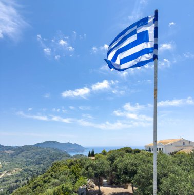 Pelekas 'ta, Korfu' da, yemyeşil tepelerde, mavi İyon Denizi 'nde ve mavi yaz gökyüzünde Yunan bayrağı dalgalanır.