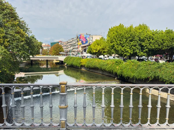 Trikala, Yunanistan, 26 Ağustos 2025. Şehir merkezinde alçak Weir ve Köprüsü olan şehir nehir kanalı, yeşil ağaçlar ve binalar, güneşli yaz günü