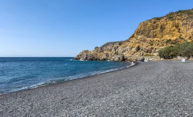 Yunanistan 'ın Chios adasındaki Mavra Volia Plajı' nda kara çakıl taşı sahili. Engebeli kayalıkların altında hafif dalgalar, mavi gökyüzü, güneşli yaz günü.
