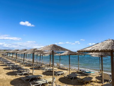 Artemida, Attica, Yunanistan 'da kumlu sahillerde saman şemsiyeler altında boş plaj salları, gün ortası güneşi ve açık mavi gökyüzü altında durgun Ege sularıyla karşı karşıya.