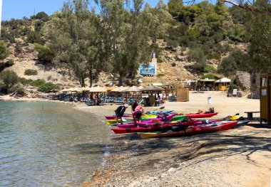Poros Adası, Yunanistan, 19 Haziran 2024. Renkli kanolar, kumlu bir sahilde insanlar saman şemsiyeler ve küçük bir sahil barı.
