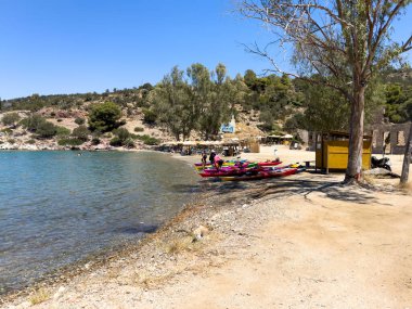 Poros Adası, Yunanistan, 19 Haziran 2024. Renkli kanolar, kumlu bir sahilde insanlar saman şemsiyeler ve küçük bir sahil barı.