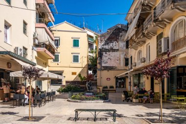 Korfu, Yunanistan, 4 Haziran 2025. Eski kasabadaki küçük bir yaya meydanı. Açık hava kafeleri, dinlenen insanlar, renkli çiçekli bir heykel ve geleneksel binalar.