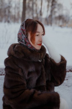 Kar yağışlı kış gününde fularlı, beyaz eldivenli kürklü kadın. Geleneksel eşarp takan ve dışarıda zarif poz veren beyaz tüylü eldiven giyen kadın. Kar zemini ve kürk manto doğal ışıkla sıcak kış atmosferi yaratır..
