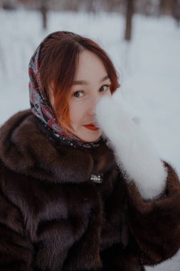 Kar kışında kürklü beyaz eldivenle yüzünü kaplayan gülümseyen kadın. Kadın, kar ve ağaçlarla çevrili, kürk manto ve desenli eşarpla dışarıda şakacı bir şekilde poz veriyor. Kırmızı ruj yumuşak doğal ışığa renk kontrastı ekler.