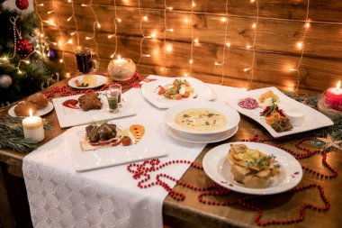 Birden fazla yemek masası, mumlar, kırmızı boncuklar ve sıcacık bokeh lambaları. Çorba, salata, ızgara et, ekmek ve sıcak şarapla hazırlanmış şenlikli Noel sofrası. Parlayan ışıklar ve hoş dekorasyonlarla aydınlatılmış..