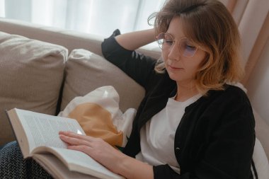 Yumuşak gün ışığı altında hafifçe yaslanmış kitap okuyan bir kadın, el destekleyici baş, hafif yansıma gösteren gözlük, sıcak yastıklar, rahat nötr ev ortamı.
