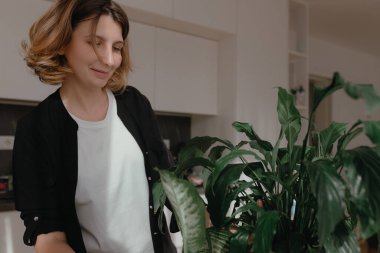Uzun bitki yapraklarını düzenlerken gülümseyen kadın, modern, parlak mutfak alanının yakınındaki kökleri aydınlatırken parlak yaprakları vurguluyor, rahat ve sıcak bir ev bitkisi bakım anı yaratıyor..
